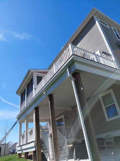 Multi-story residential house with white railings, covered porch, and large support columns under clear blue sky