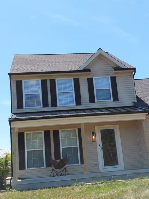 Two-story beige house with black shutters, front porch with chair, and dormer window under clear blue sky