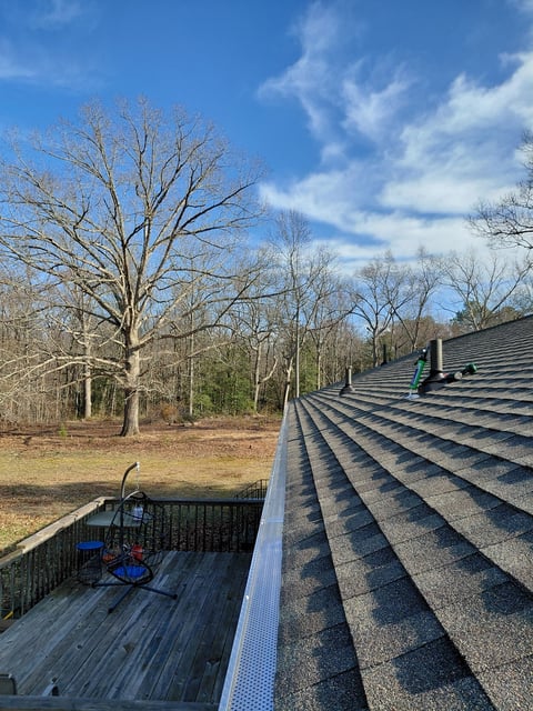 Roofer working on a dark shingled roof with a wooden deck and bare trees visible in the wooded background