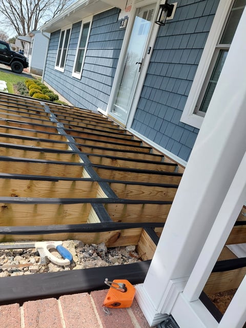 Construction view of house deck framing with wooden joists, blue house siding, and an orange power tool on brick foundation