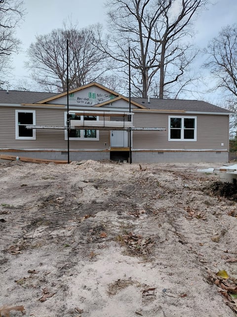 New modular home under construction on sandy lot with bare trees in background