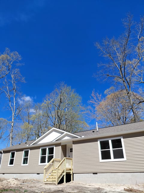 Single-story beige mobile home with white trim and yellow steps under a clear blue sky with bare trees