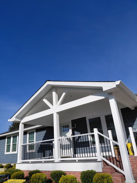 Modern home with white porch and pergola featuring X-pattern beams, black metal railings, brick foundation, and clear blue sky
